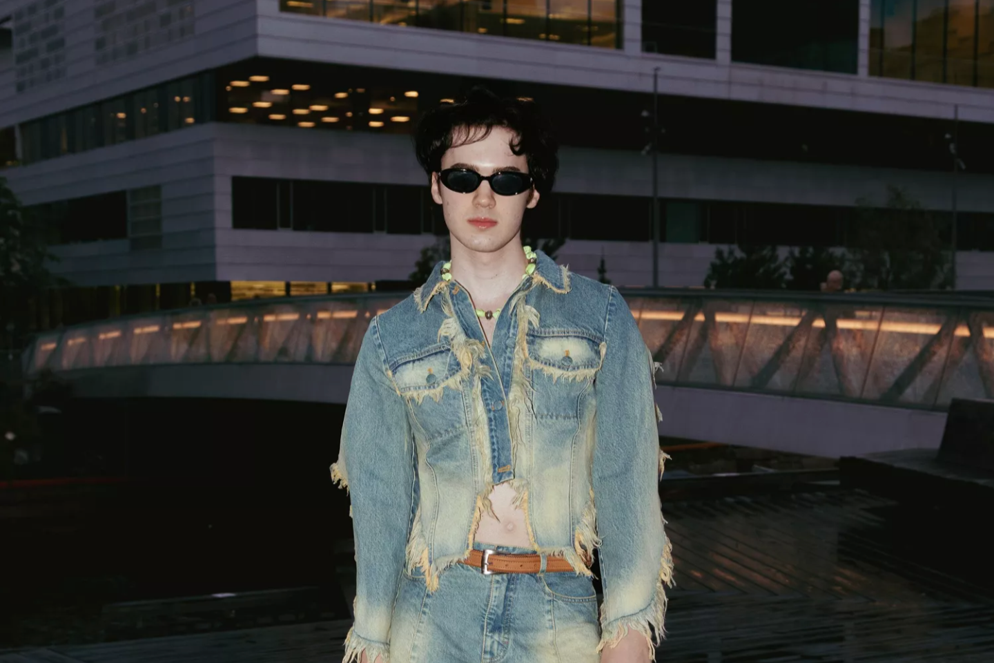 The founder of Heim Studios wearing a denim outfit with a fringe skirt in front of a building at night.